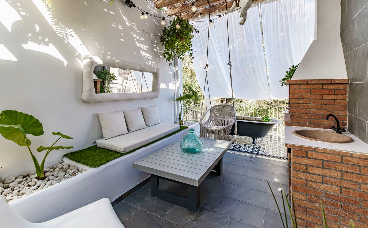 Terrasse blanche avec canapé intégré, balançoire macramé, évier en brique et baignoire noire sous des voiles.
