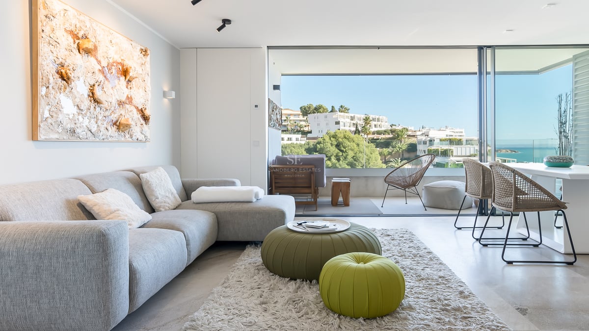 Salón moderno y luminoso con sofá gris y pufs verdes, con ventanales de suelo a techo y vistas al mar.
