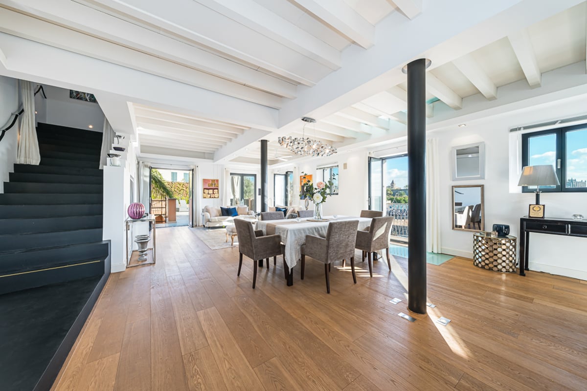 Pièce lumineuse à plan ouvert avec parquet, plafond à poutres blanches, coin repas et escalier noir sur la gauche.