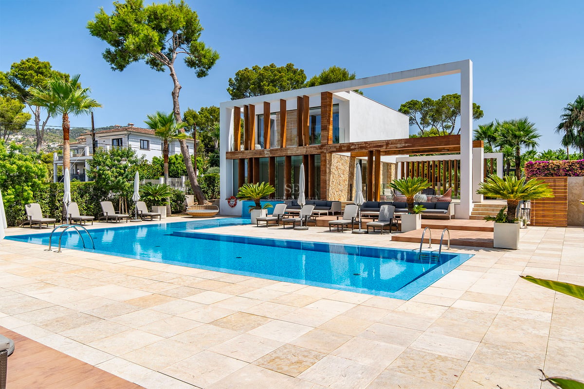 Grande piscine bleue entourée d'une terrasse en pierre claire et d'une villa blanche moderne avec accents en bois.