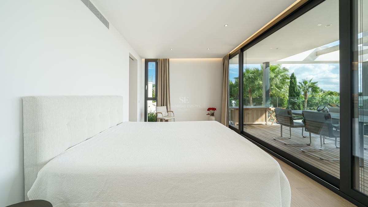 Chambre de maître minimaliste blanche avec baies vitrées ouvrant sur une terrasse en bois ensoleillée et des palmiers.