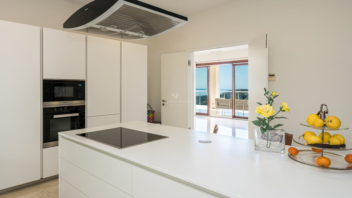 Cuisine moderne blanche et lumineuse avec îlot central, appareils Miele intégrés et vue sur la terrasse et la mer.