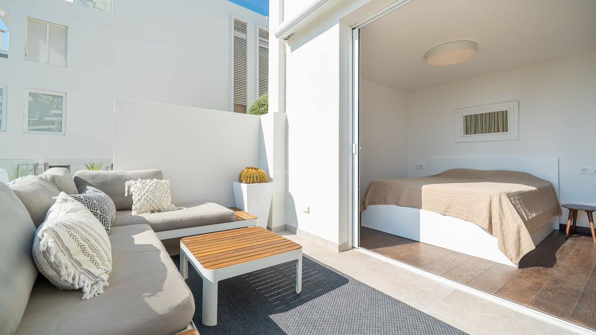 Chambre moderne s'ouvrant sur une terrasse ensoleillée avec canapé lounge, table en teck et murs blancs épurés.