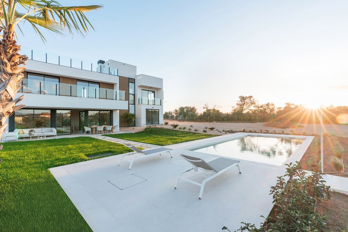 Moderne weiße Villa mit Pool, Sonnenliegen und grünem Rasen unter einem goldenen Sonnenuntergang.