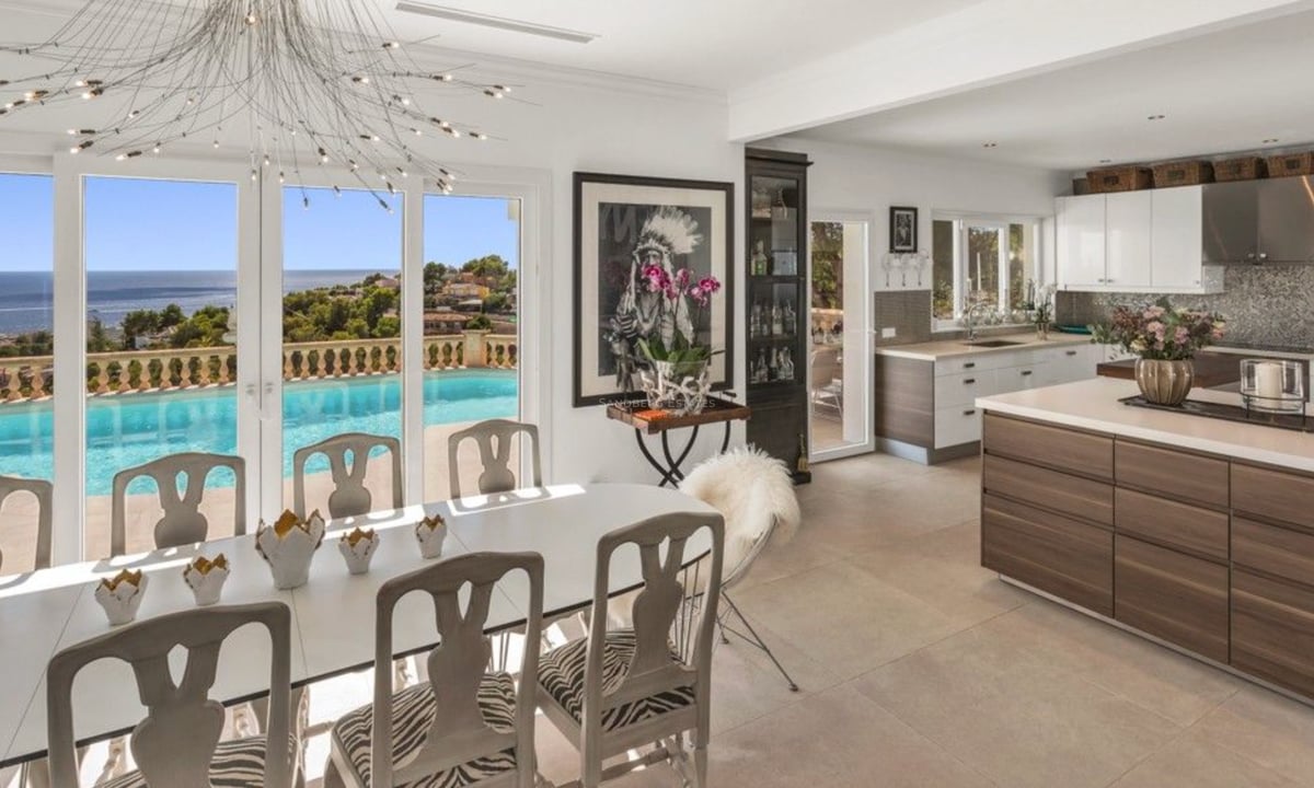 Salle à manger et cuisine modernes avec meubles blancs, donnant sur une piscine à débordement et la mer Méditerranée.