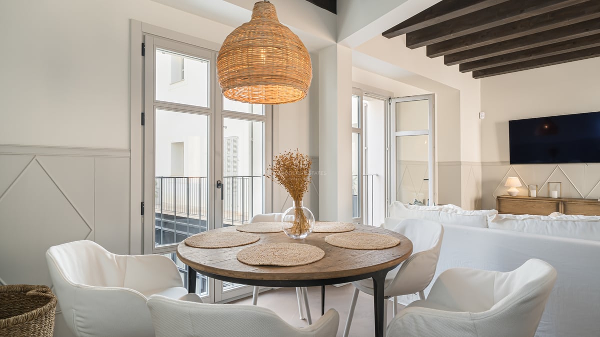Mesa de comedor redonda de madera, sillas blancas, lámpara de mimbre y vigas oscuras en un salón luminoso.