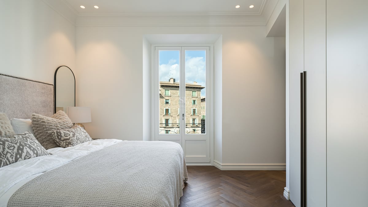 Bright bedroom with dark wood herringbone floors, white built-in wardrobes, and a window overlooking historic buildings.