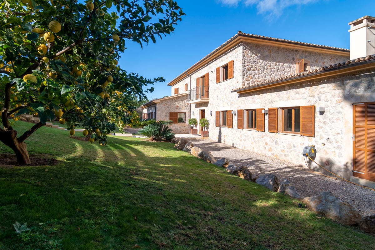 Extérieur d'une villa en pierre naturelle avec volets en bois, pelouse et citronniers sous un ciel bleu.