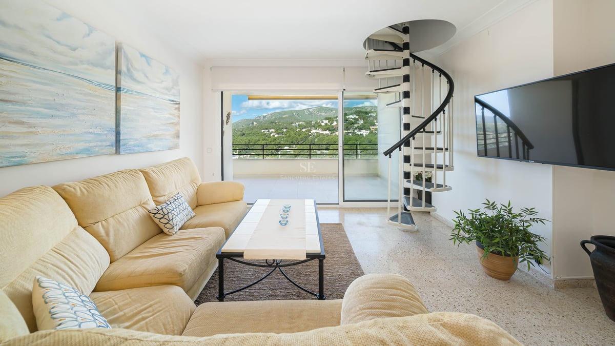 Salon lumineux avec canapé crème, escalier en colimaçon noir et blanc et balcon avec vue sur la montagne.