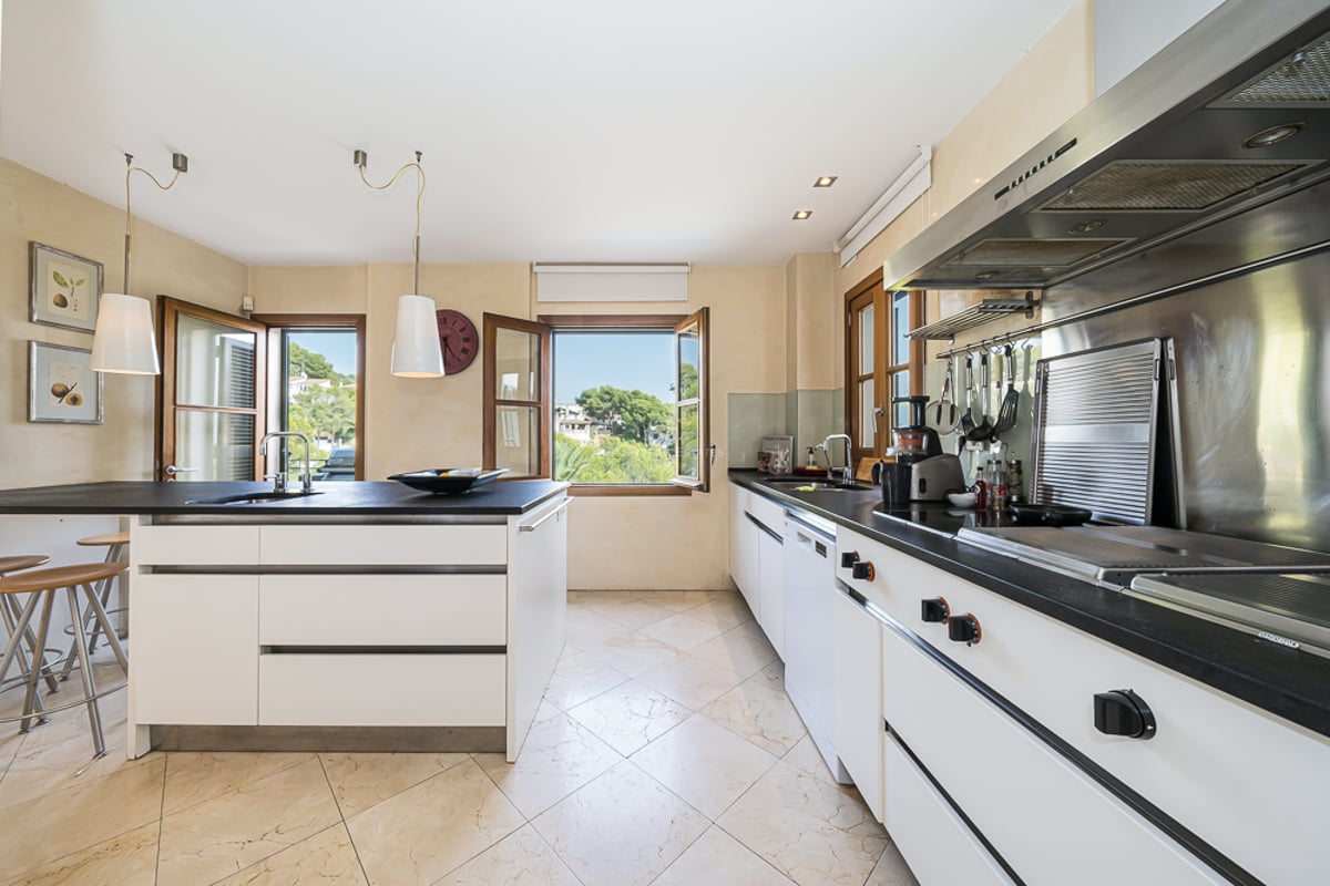 Cuisine contemporaine blanche avec plans de travail sombres, sol en carrelage et fenêtres donnant sur la verdure.