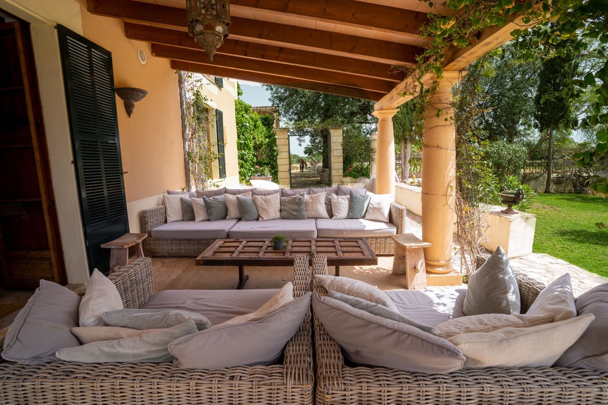 Terrasse couverte avec canapés en osier, poutres en bois apparentes et colonnes en pierre donnant sur le jardin.