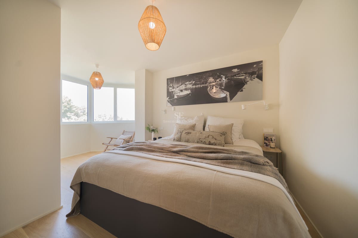 Chambre lumineuse avec un grand lit, des suspensions en osier et une grande fenêtre latérale.