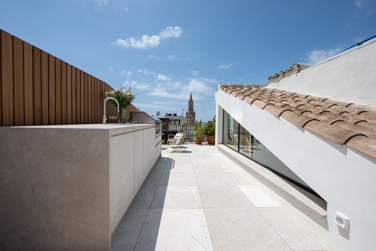 Terraza moderna en la azotea con cocina exterior, suelo de piedra y vistas a una catedral bajo un cielo azul.