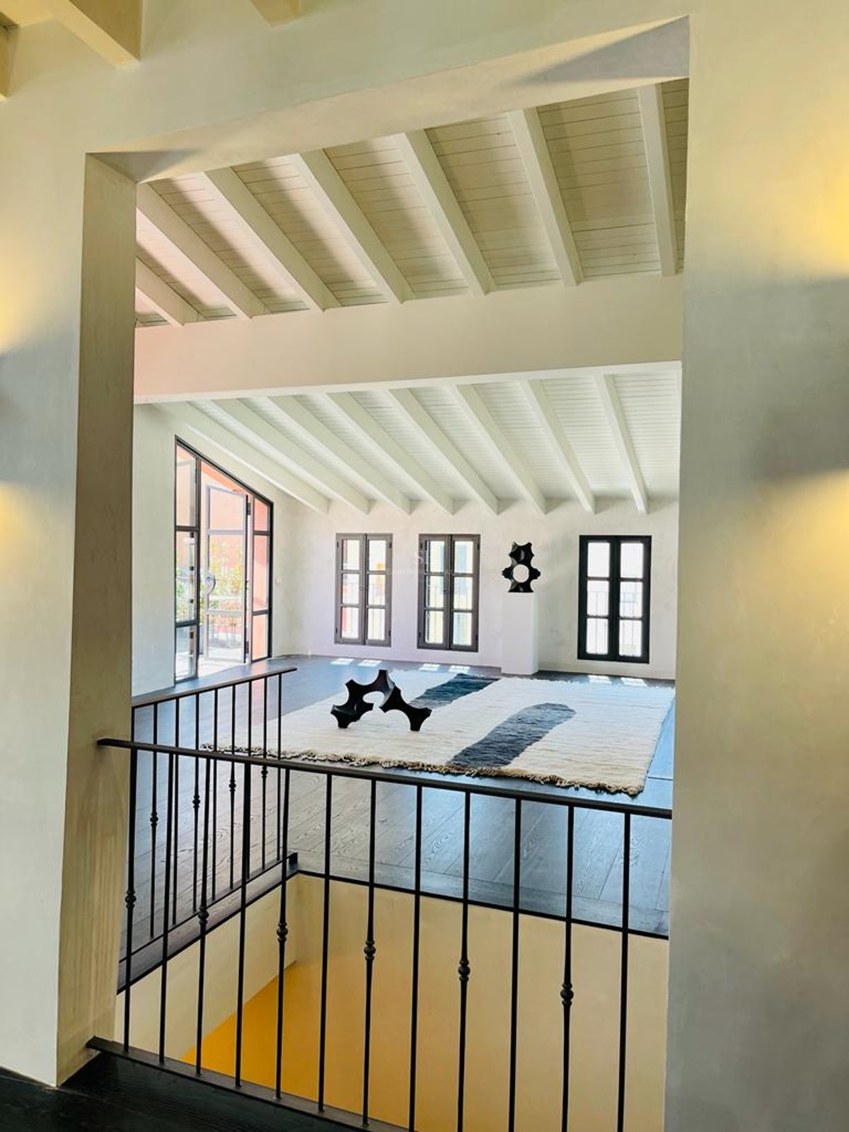 Vue intérieure depuis une mezzanine montrant un salon avec parquet sombre, poutres blanches et fenêtres industrielles.