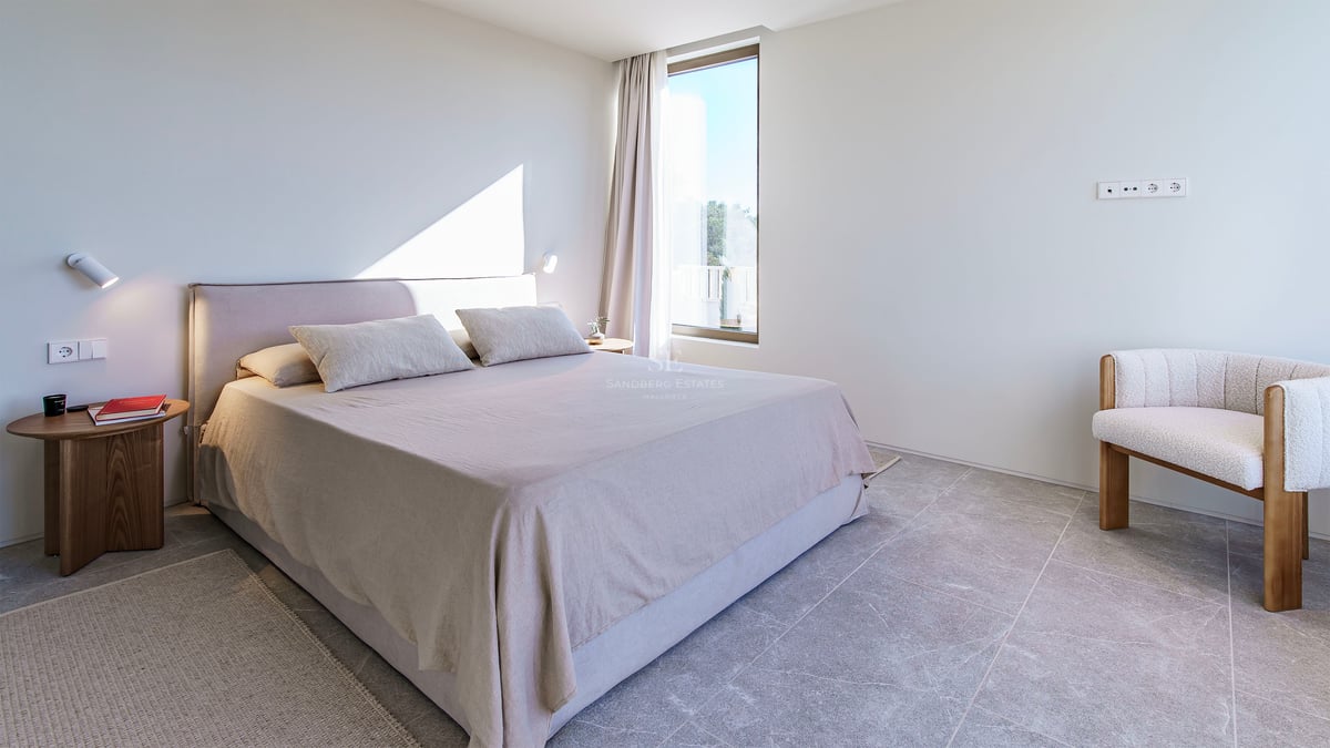 Chambre lumineuse avec carrelage en pierre grise, lit rembourré beige, table de chevet en bois et fauteuil design.