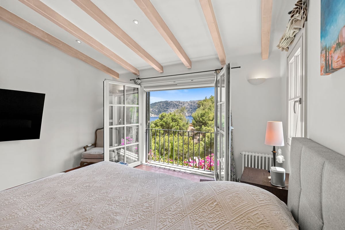 Chambre lumineuse avec poutres en bois et balcon ouvert donnant sur la mer et les collines.