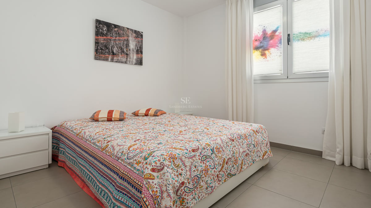Chambre moderne aux murs blancs, sol en carrelage gris, lit double avec couvre-lit coloré et rideaux blancs.