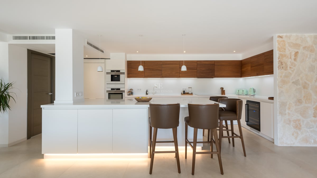 Cuisine moderne avec îlot blanc, tabourets en cuir, placards en noyer et mur en pierre naturelle.