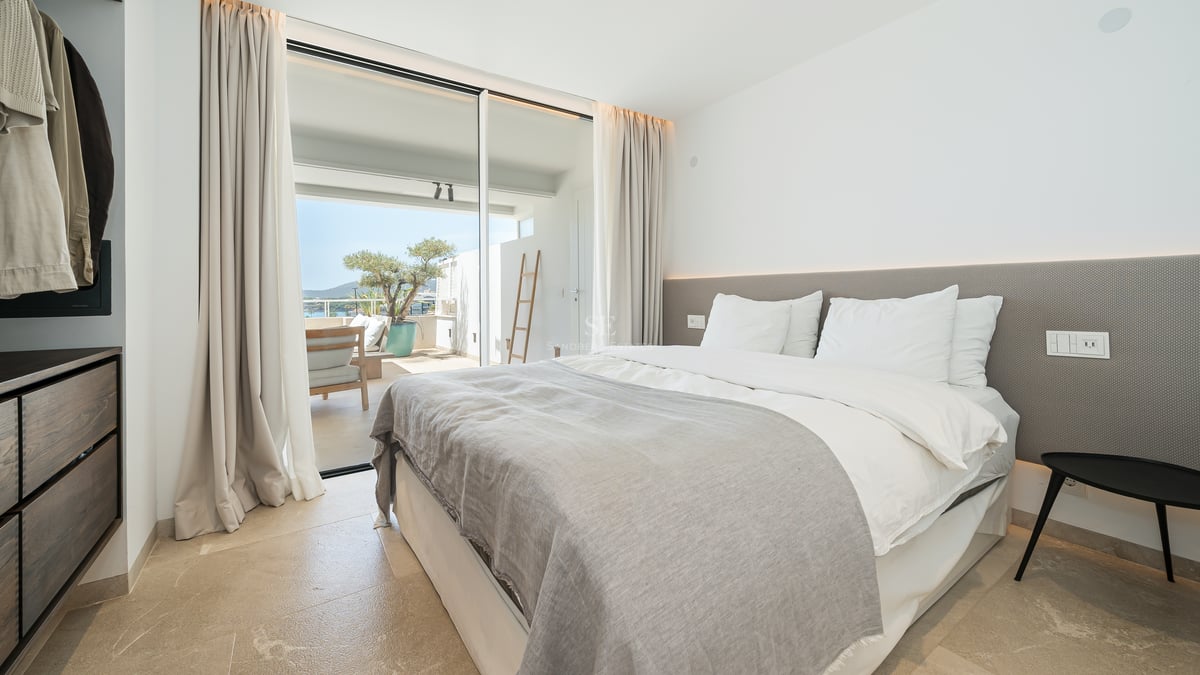 Chambre lumineuse avec grand lit, sol en pierre et baies vitrées ouvrant sur une terrasse ensoleillée.