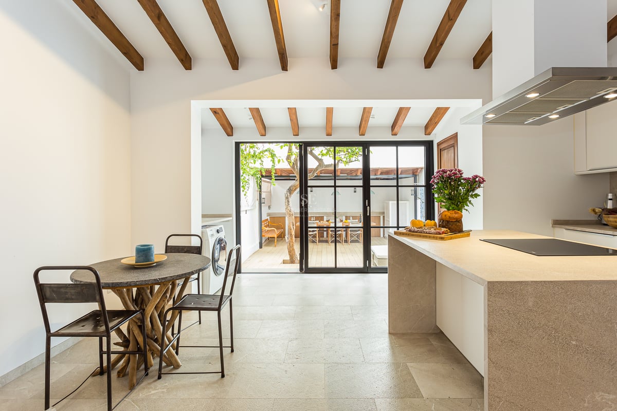 Cuisine lumineuse avec poutres en bois, îlot en pierre et portes vitrées s'ouvrant sur un patio avec un arbre.