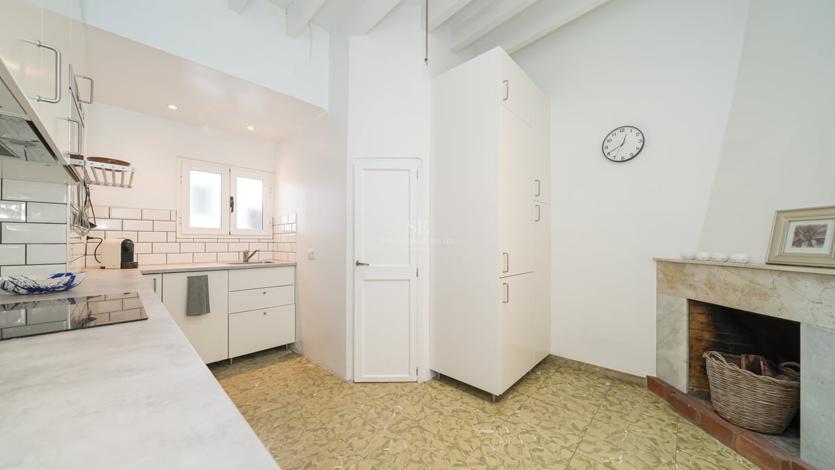 Cuisine blanche lumineuse avec cheminée en marbre, plafond à poutres et carrelage à motifs.