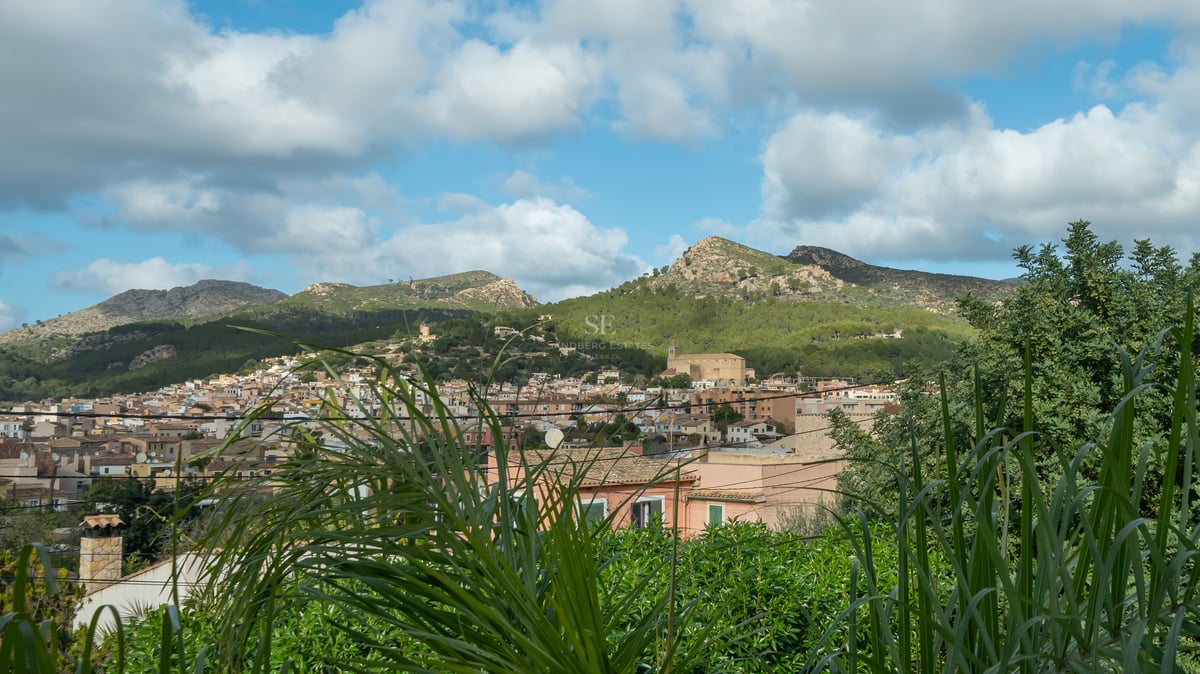 Vue d'un village méditerranéen aux toits de terre cuite entouré de montagnes verdoyantes sous un ciel nuageux.