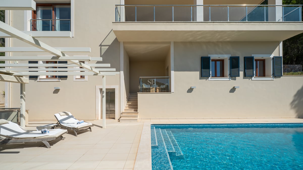 Piscine de villa de luxe avec mosaïque et chaises longues blanches sur une terrasse en pierre sous une pergola.