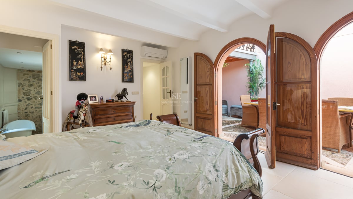 Chambre avec portes en bois voûtées ouvrant sur un patio ensoleillé, comprenant une salle de bain avec mur en pierre.