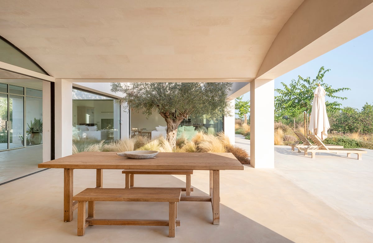 A wooden dining table on a covered sandstone terrace next to an olive tree and sun loungers.