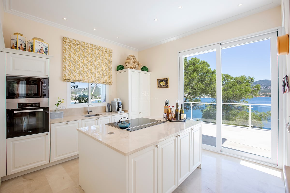 Cuisine moderne crème avec îlot central et grandes baies vitrées donnant sur la mer et des pins.