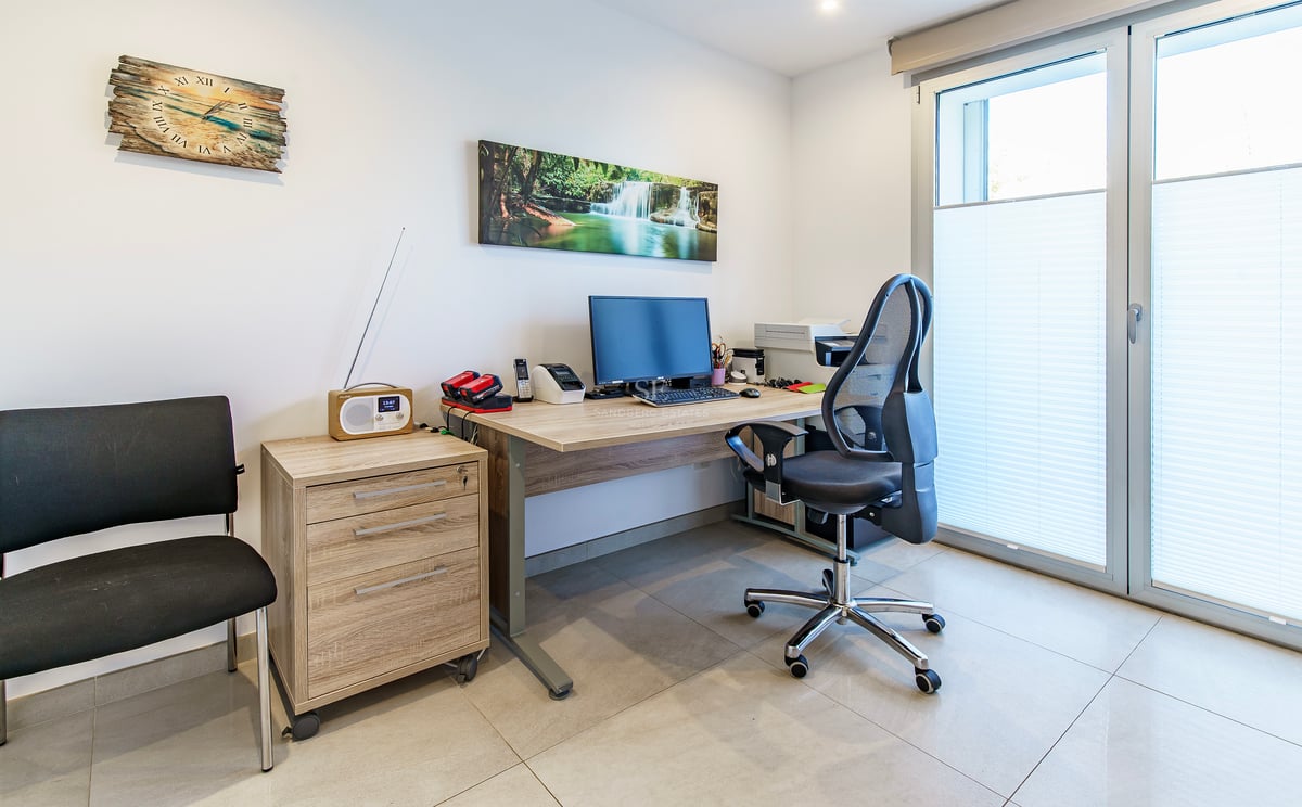 Bureau avec bureau en bois, chaise ergonomique, ordinateur et grande porte-fenêtre avec stores.