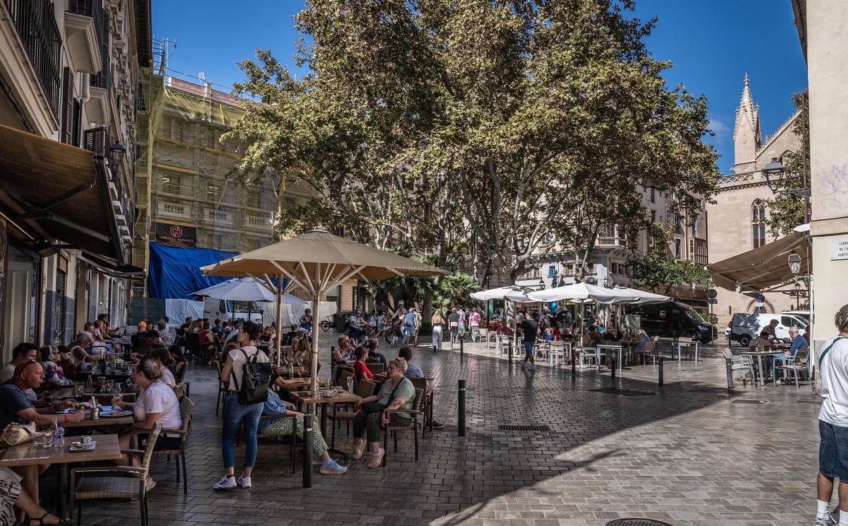 Des gens assis à des tables de café en plein air sous des parasols sur une place pavée avec des arbres et des bâtiments.