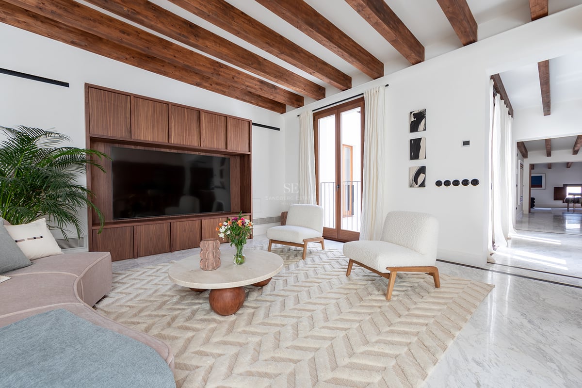 Salón luminoso con vigas de madera oscura, suelo de mármol, sillones bouclé blancos y mueble de televisión a medida.