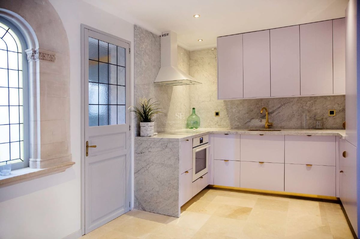 Cocina contemporánea con armarios blancos, salpicadero de mármol, grifo dorado y una ventana de piedra arqueada.