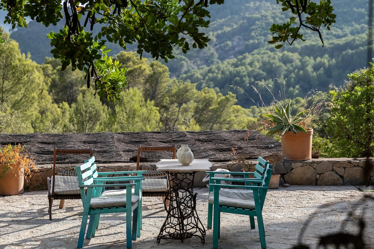Terrasse en pierre avec chaises turquoise et table vintage surplombant une forêt verdoyante et des montagnes.
