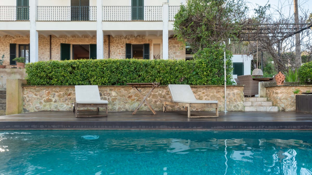 Piscine turquoise avec chaises longues en bois devant une villa en pierre aux volets sombres.