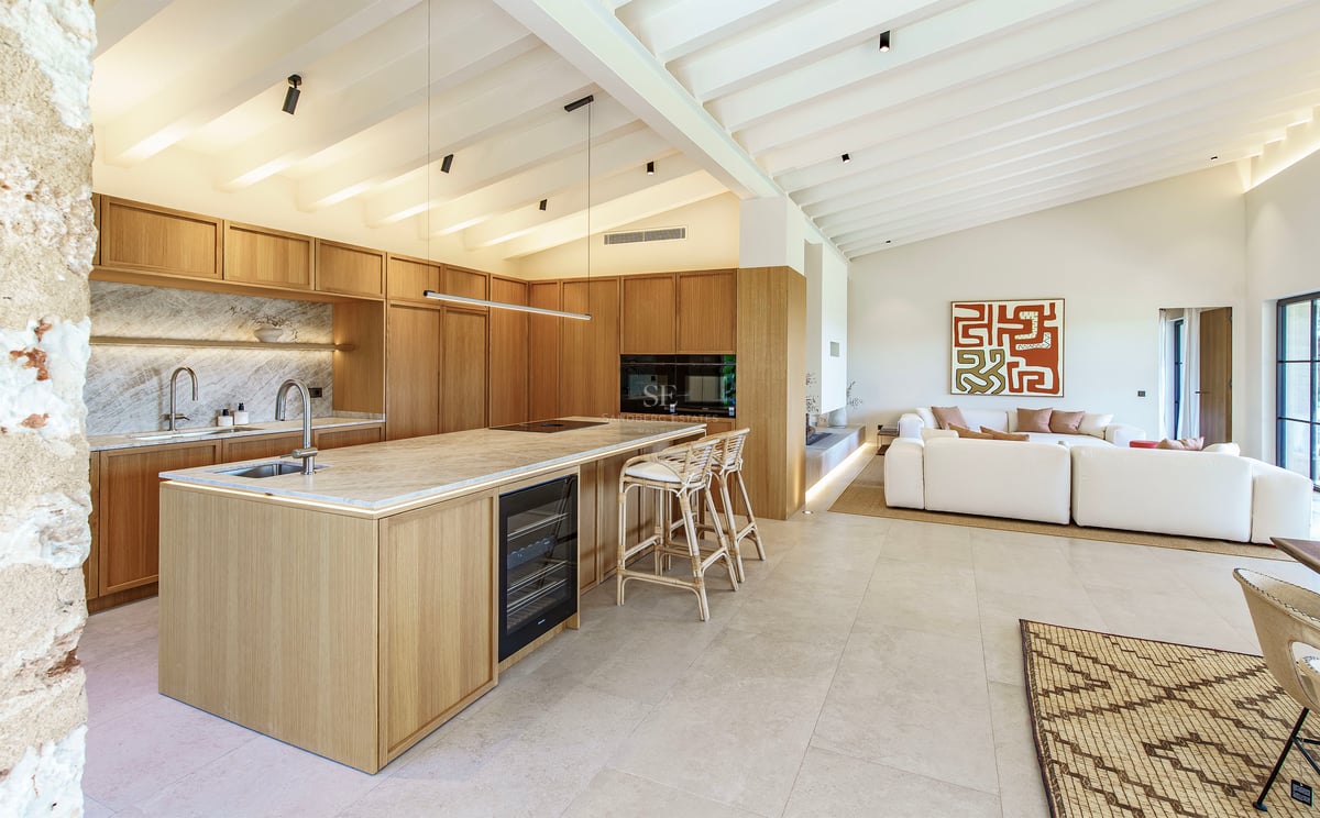Cocina moderna de planta abierta con muebles de madera clara, isla de mármol y techo de vigas blancas.