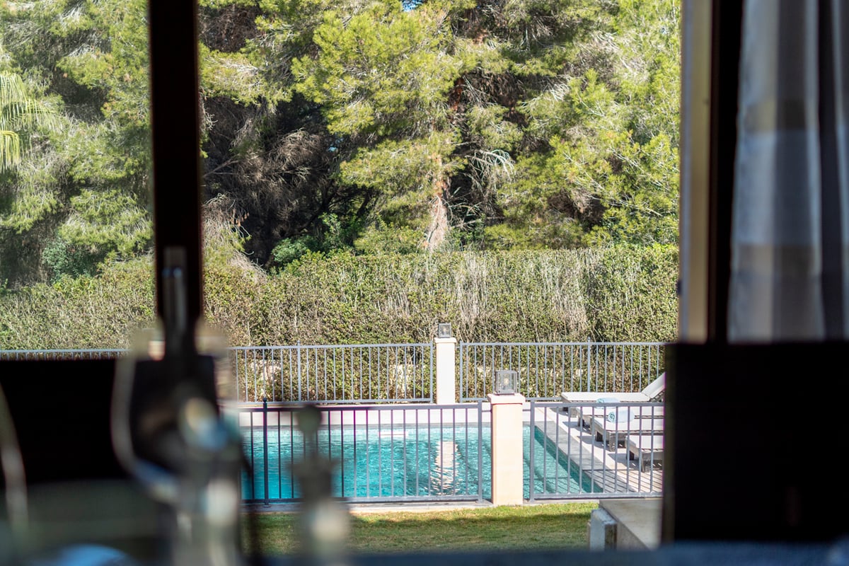 Vue par une fenêtre d'une piscine turquoise clôturée entourée de transats et de grands pins.