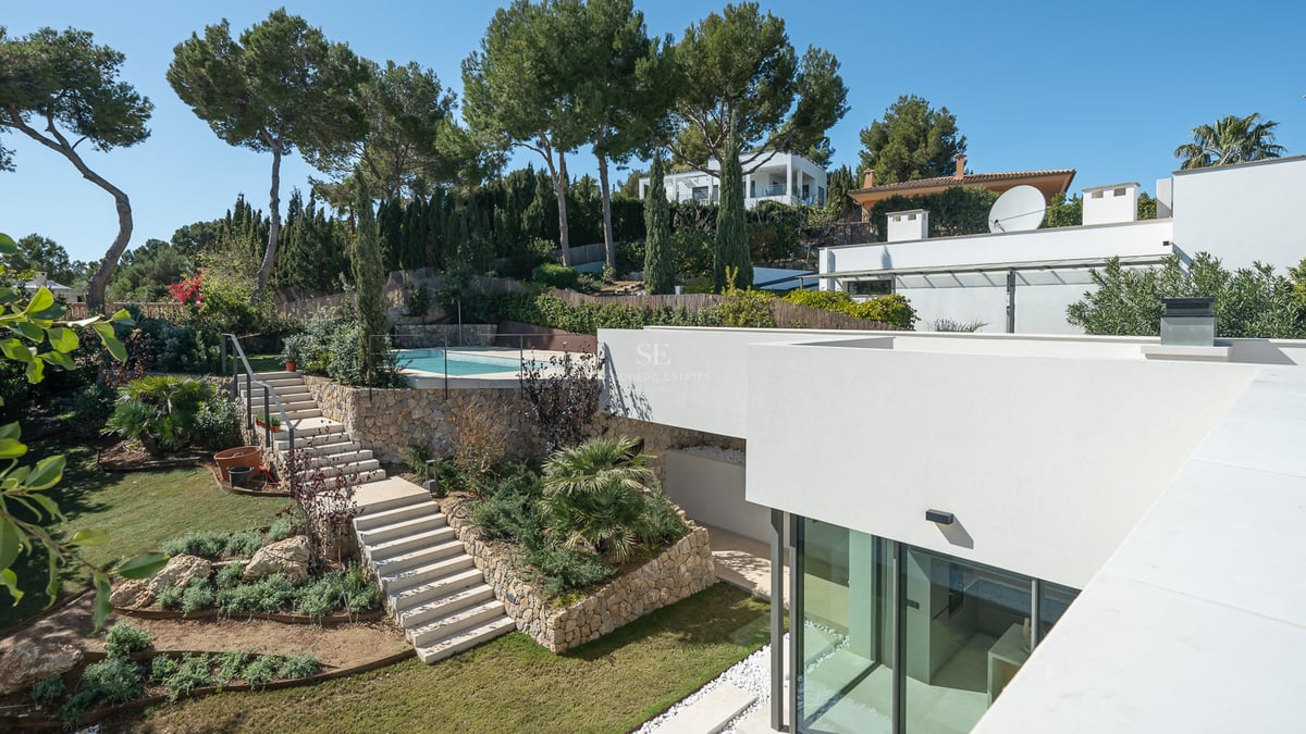 Villa blanche moderne avec jardin en terrasses, escaliers en pierre et piscine entourée de pins sous un ciel bleu.