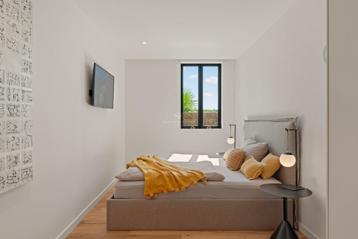 Chambre lumineuse avec lit beige, plaid jaune, lampes design et fenêtre donnant sur un jardin.