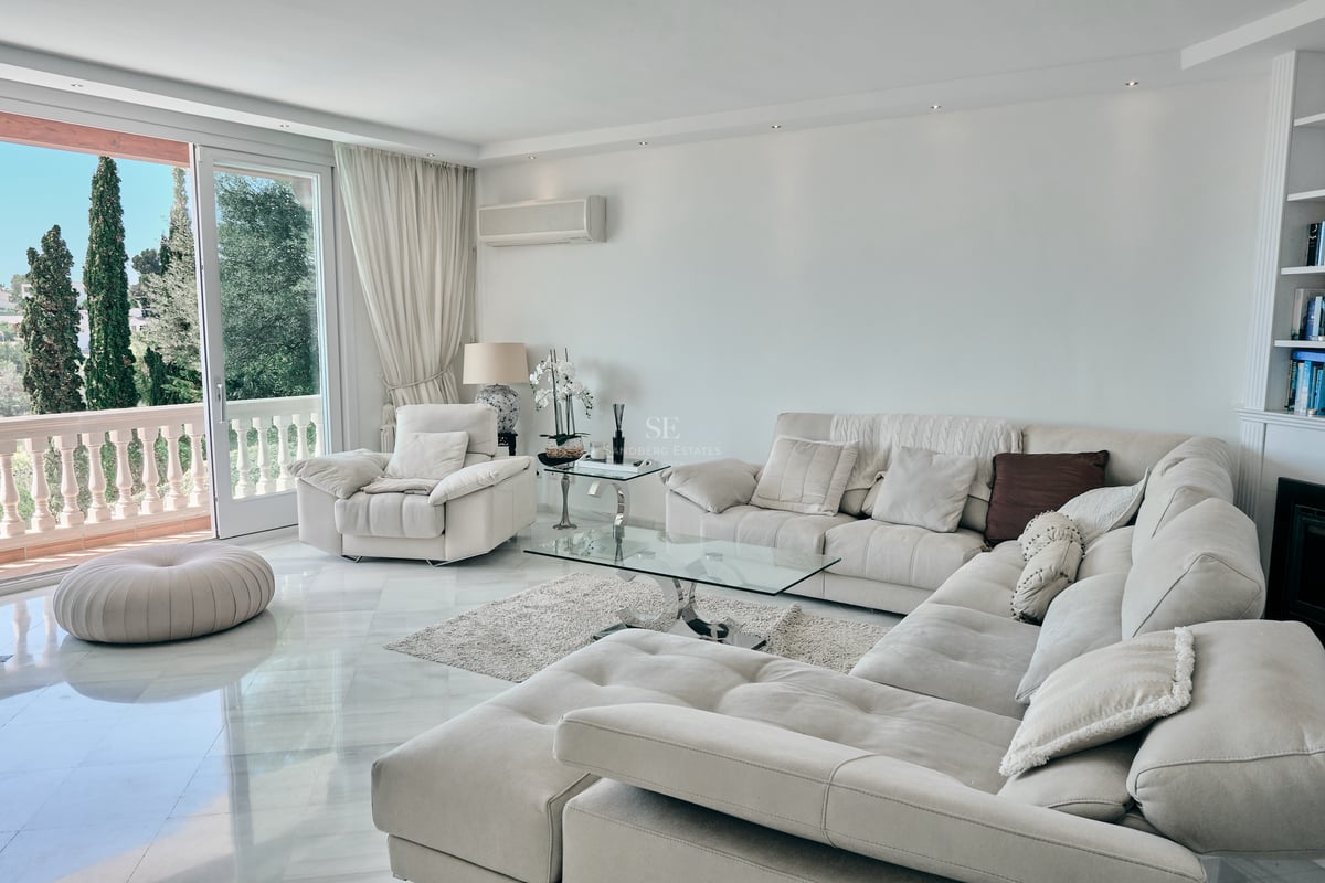 Salon spacieux avec canapé d'angle beige, sol en marbre blanc et vue sur jardin méditerranéen.