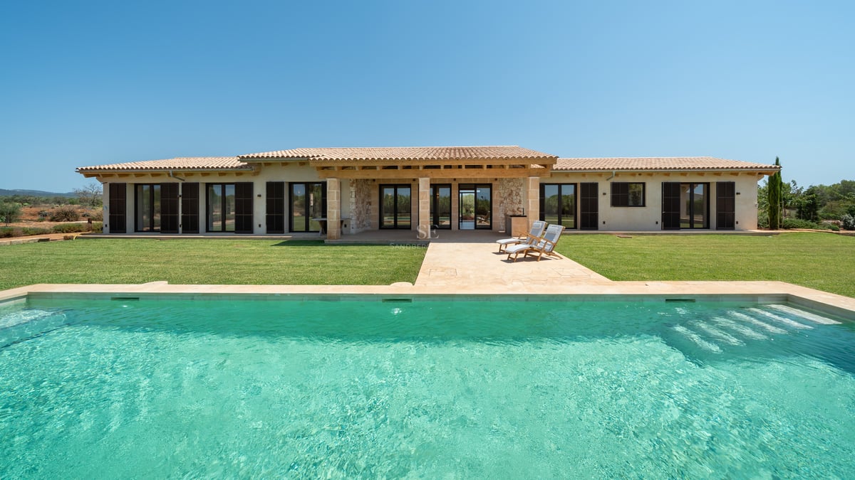 Piscina rectangular turquesa frente a una villa moderna de piedra de una planta con persianas oscuras y jardín.