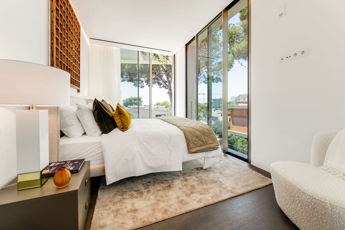 Chambre contemporaine lumineuse avec lit blanc, parquet foncé, fauteuil bouclé et parois vitrées.