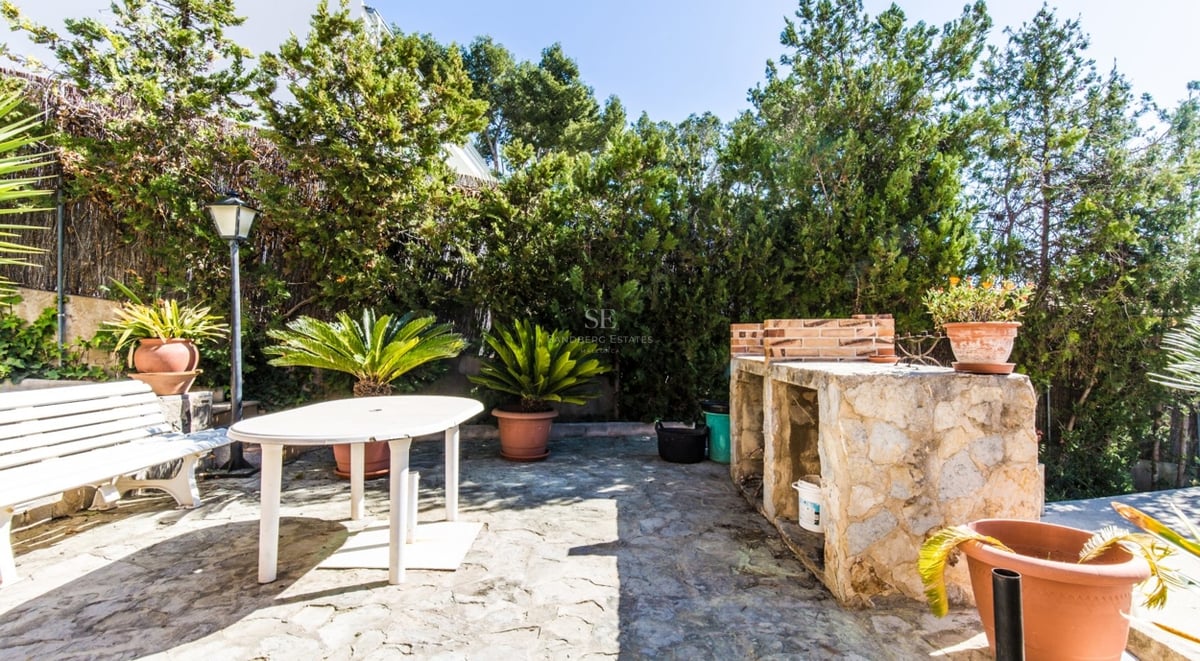 Patio pavé avec barbecue en pierre, table et banc blancs, entouré de haies vertes et de plantes en pots.
