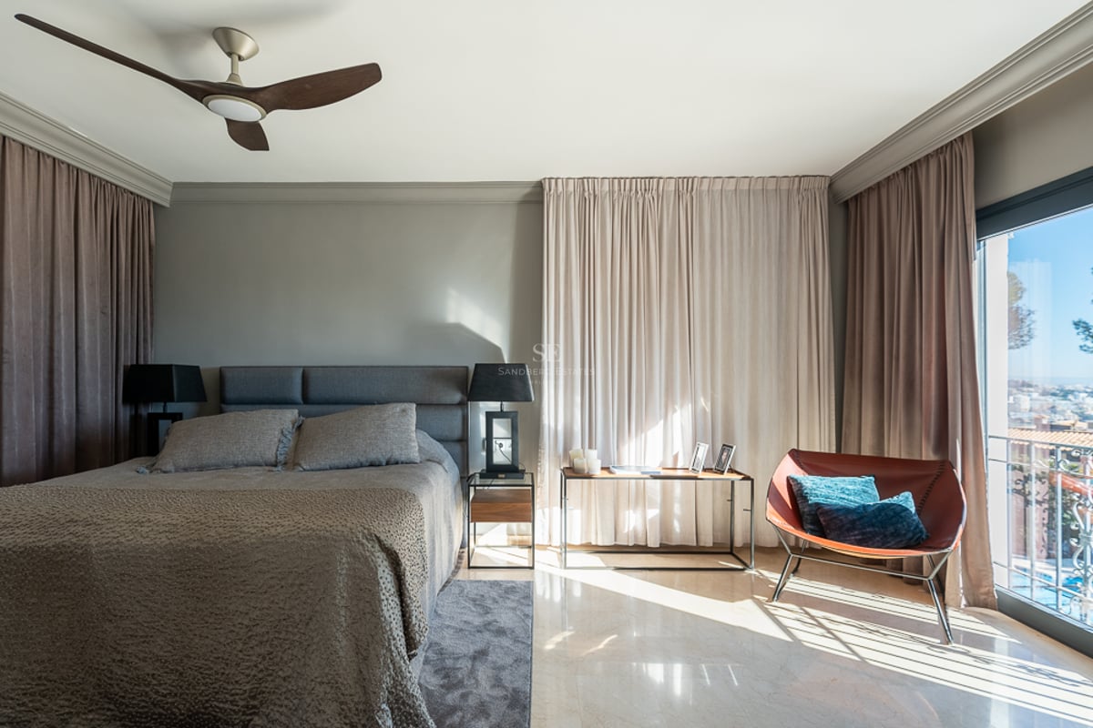 Chambre moderne avec lit tapissé gris, sol en marbre, fauteuil en cuir et balcon offrant une vue sur la côte.