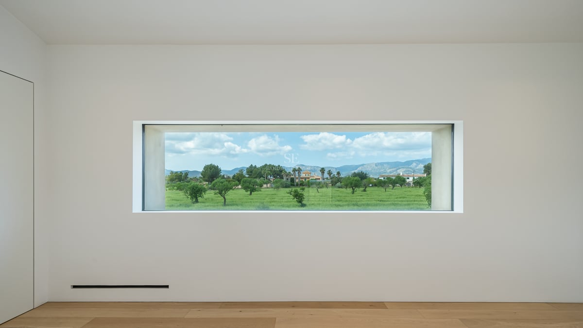 Une fenêtre rectangulaire cadre une vue sur des champs verts et des montagnes sous un ciel bleu dans une pièce sobre.