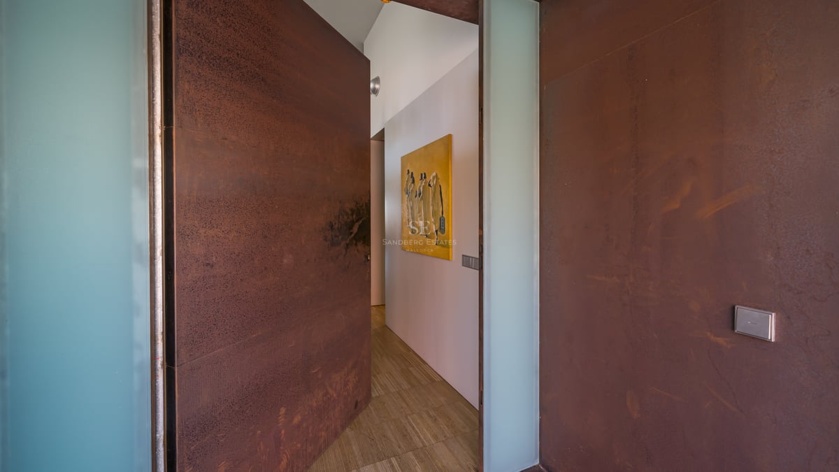 Grande porte en acier Corten s'ouvrant sur un couloir minimaliste avec murs blancs et sol en pierre.