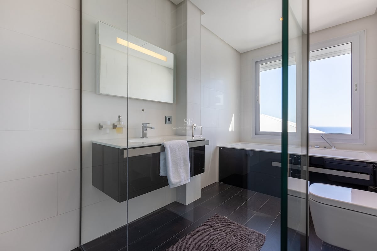 Salle de bain moderne avec murs carrelés blancs, sol sombre, vanité suspendue et baignoire sous une grande fenêtre.