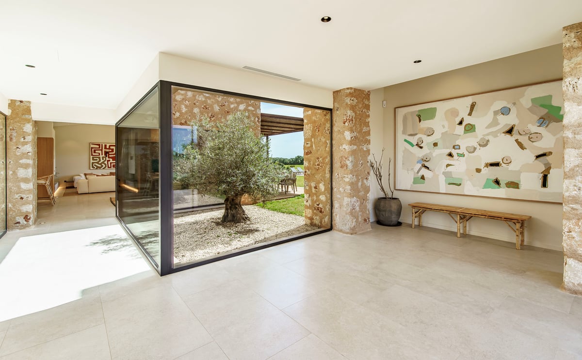 Pasillo moderno con grandes ventanales que muestran un patio interior con un olivo y paredes de piedra natural.