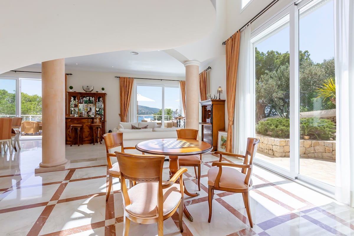 Spacieux salon avec sol en marbre, table en bois, colonnes et grandes fenêtres donnant sur un jardin et la mer.